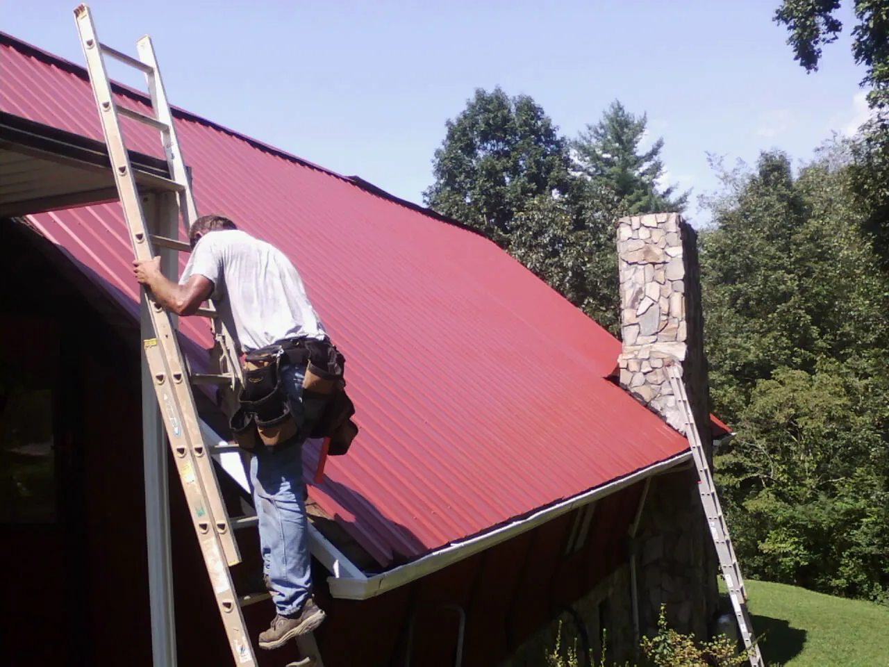 Roofer on a ladder beside a red metal roof and stone chimney — Soffit & Fascia Repair by Roofing Brothers in Live Oak